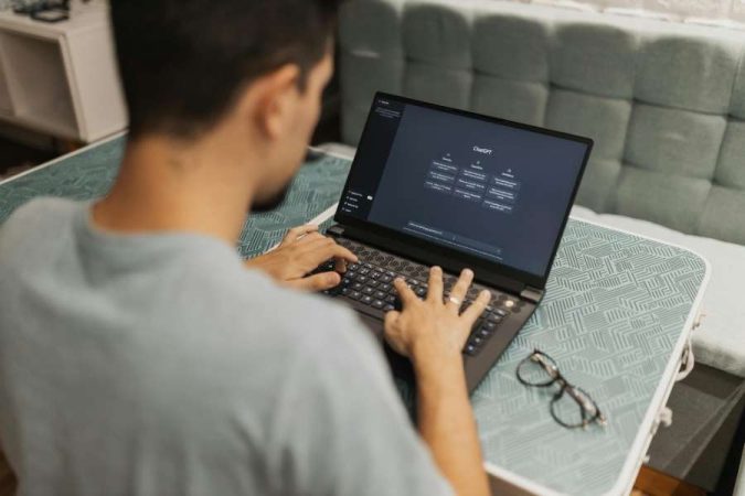 Vue de dessus d'un homme utilisant l'interface de ChatGPT sur son ordinateur portable posé sur une petite table de salon.