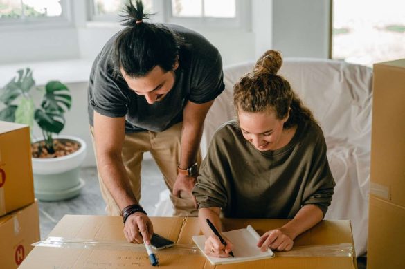 Un couple sourit en remplissant une liste changement d'adresse déménagement à imprimer posée sur un carton de déménagement.